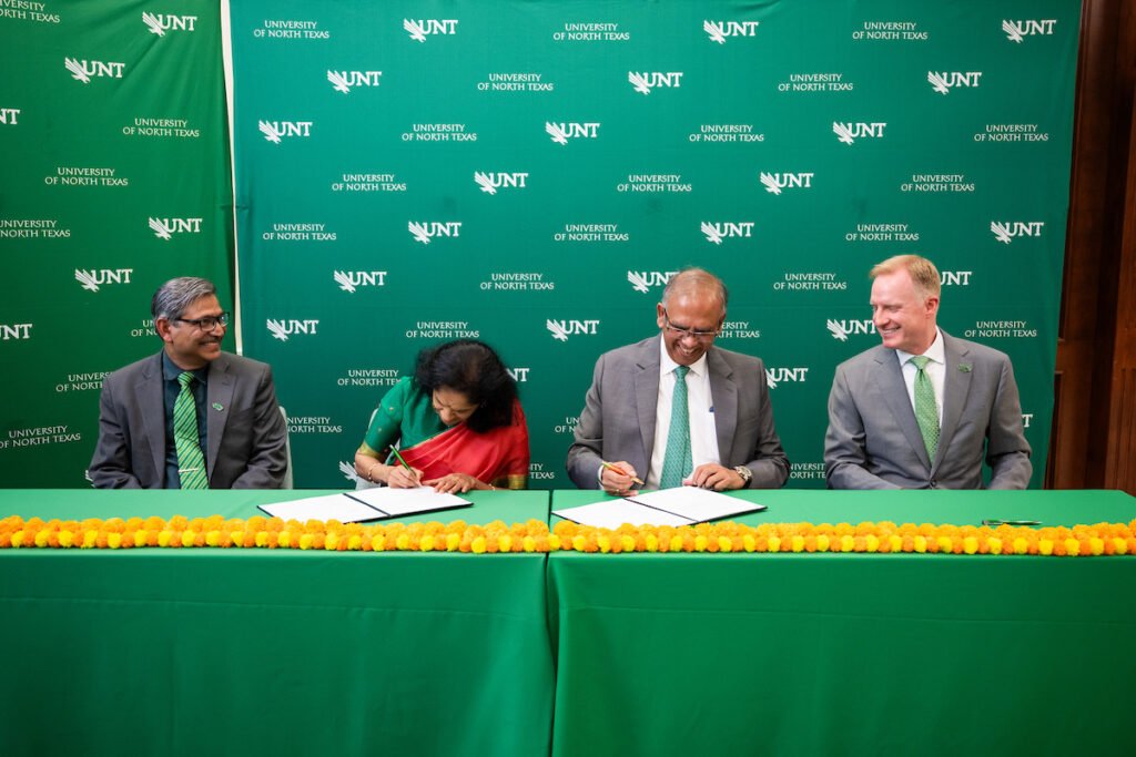 Four individuals in formal attire sign documents at a green table adorned with orange flowers, seated before a backdrop featuring the University of North Texas logo.